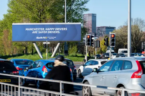 A Kwik Fit roadside billboard reading ‘Manchester, happy drivers are safer drivers’ above queuing traffic