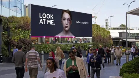A large Women’s Aid billboard reading ‘Look at me. We can stop it.’ showing a woman with a bruised face, above passers-by
