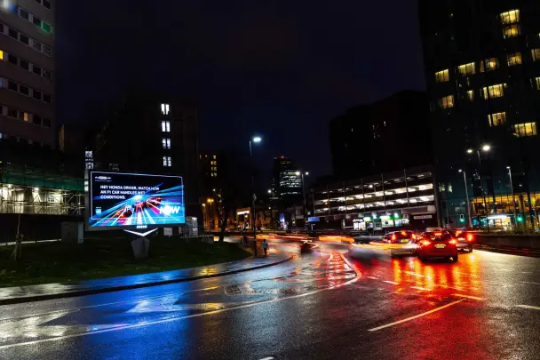 A roadside NOW billboard at night telling passing Honda drivers how an F1 car handles wet conditions, beside a rain-soaked road