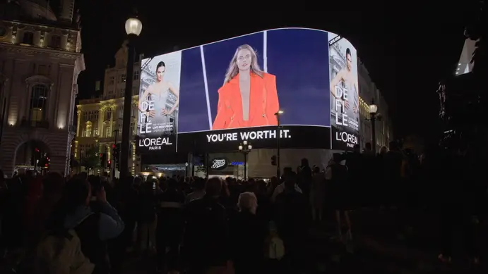 A crowd in Piccadilly Circus at night beneath the curved Piccadilly Lights screen, showing L’Oréal Paris’s Le Défilé runway show live from the Opéra de Paris
