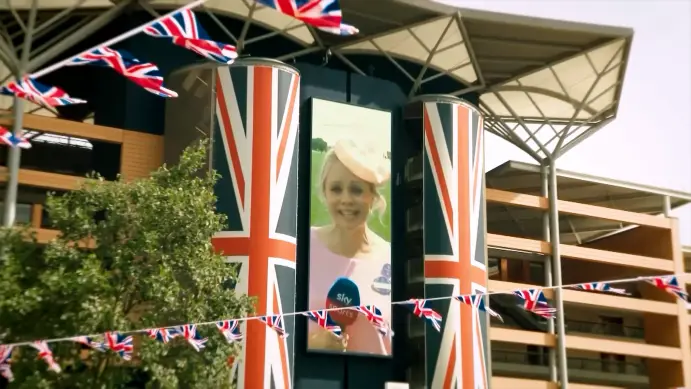 A portrait LED screen between Union Jack–wrapped pillars at Ascot, showing a Sky presenter in a pink hat