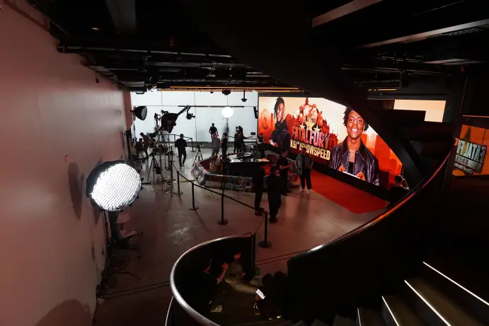 Looking down a spiral staircase onto a production set, where a large LED wall shows Fatal Fury artwork featuring IShowSpeed