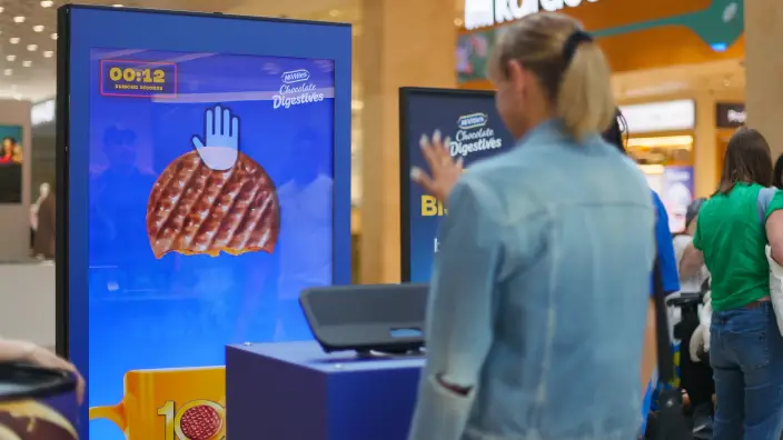 A shopper playing McVitie’s Dare to Dunk in a shopping centre, using hand gestures to dunk a biscuit on a portrait screen against a 12-second timer