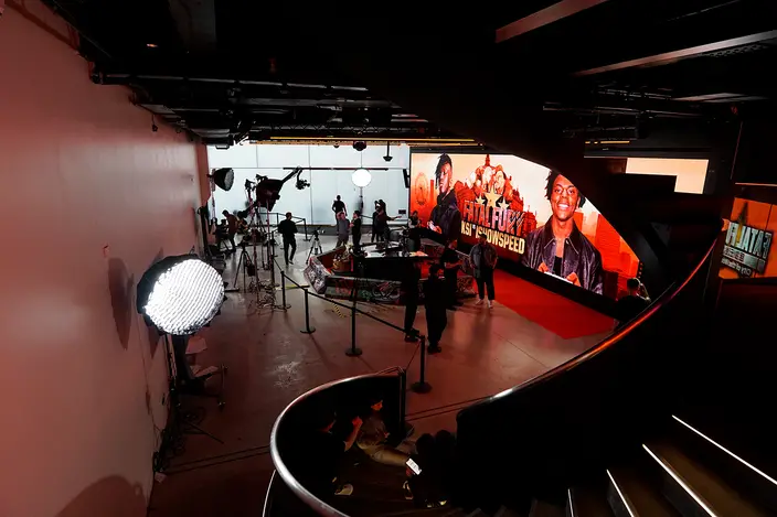 A film crew setting up lights and cameras in front of a large LED wall showing Fatal Fury artwork with KSI and IShowSpeed, seen from a spiral staircase