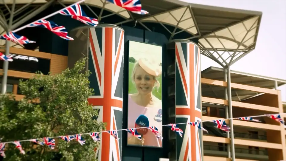 A portrait LED screen between Union Jack–wrapped pillars at Royal Ascot, showing a Sky presenter in a pink hat
